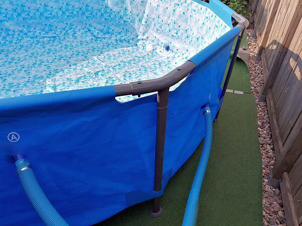 Ein kleiner Pool für den Garten - Schritt für Schritt. Hier: Detailansicht eines blauen Aufstellpools im Garten mit angeschlossenen Poolschläuchen und stabilem Rahmen – die Technik ist vorbereitet, damit dem ungetrübten Badespaß an warmen Sommertagen nichts mehr im Weg steht.