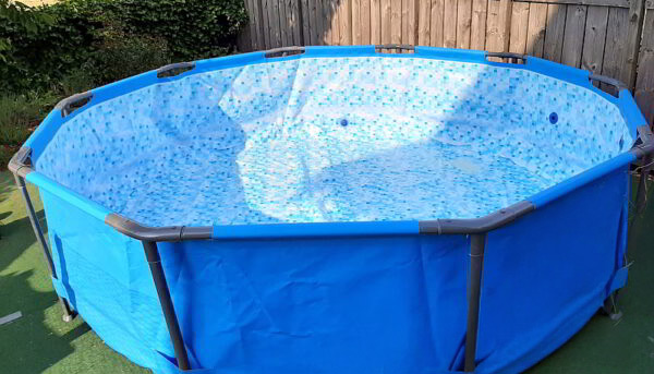 Ein Aufstellpool für den Garten. Hier zu sehen: Sechseckiger, blauer Aufstellpool im Garten, frisch aufgebaut und noch leer, mit strahlender Innenfolie – bereit für die ersten sonnigen Badetage, eingerahmt von grünem Rasen und einem Holzzaun.
