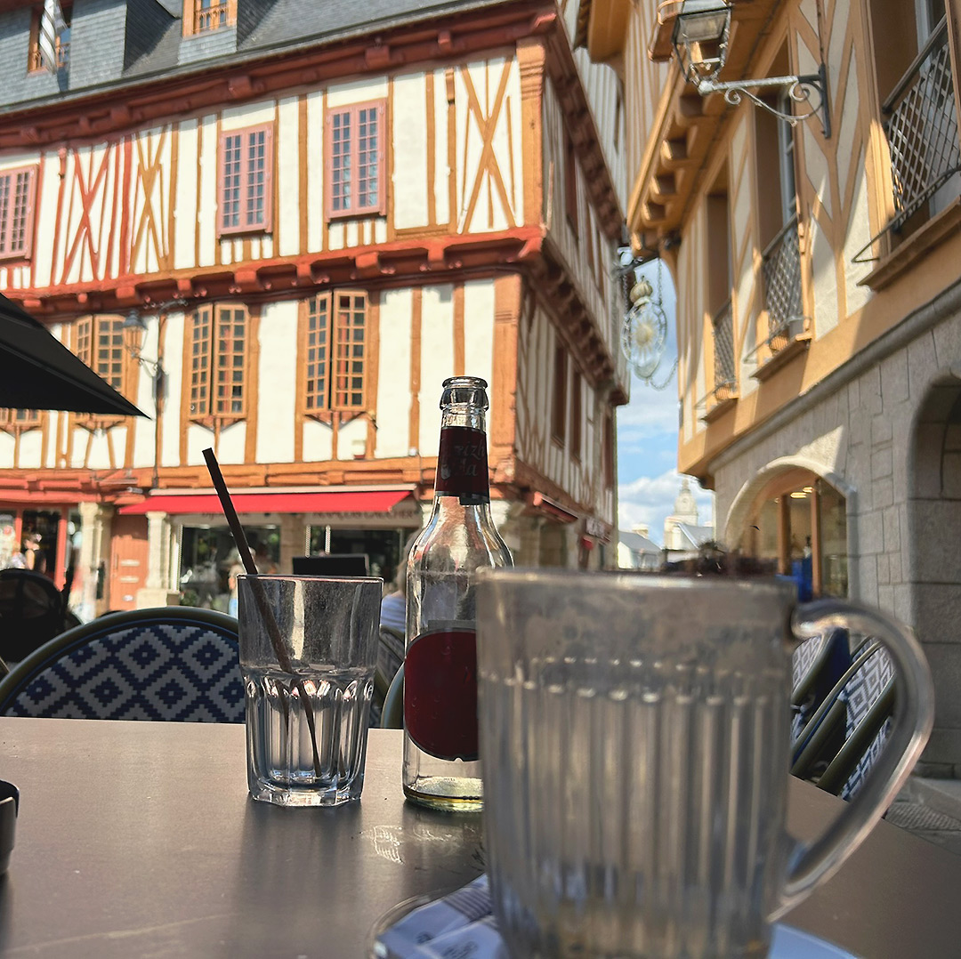 Bretagne mit Kindern - Südküste Südliche Bretagne. Ein Ausflug nach Vannes, hier ein Blick auf ein wunderschönes Fachwerkhaus - im Vordergrund ist ein Tisch eines Cafés zu sehen mit Gläsern und Flaschen.