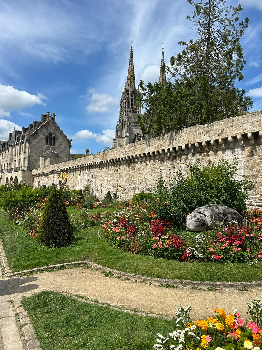 Bretagne mit Kindern - Westküste Bretagne - der Westen. Hier ein Bild der Stadtmauer von Quimper. Im Hintergrund ragen die Türme der Kathedrale auf. Im Vordergrund ist eine schöne Gartenanlage mit Sommerblumen zu sehen.
