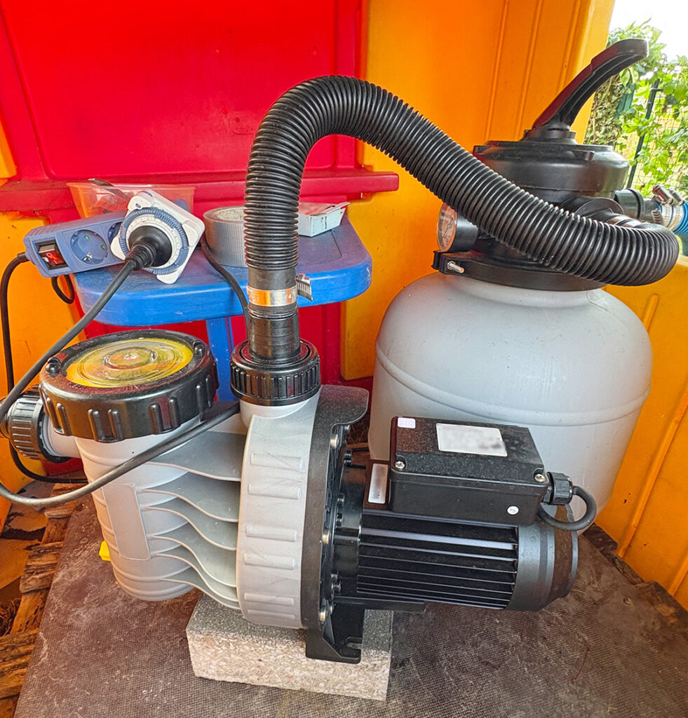 familie-entdeckt.de - hier zu sehen - eine Pool-Filteranlage bestehend aus einer Pumpe und einem grauen Sandfilter steht in einem bunten Geräteschrank. Die Komponenten sind mit einem schwarzen Wellschlauch verbunden. Im Hintergrund befinden sich eine Zeitschaltuhr und weiteres Zubehör zur Steuerung der Pooltechnik.